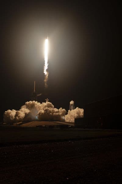 Noční start rakety Falcon 9 z Kennedyho vesmírného střediska na Floridě — první stupeň stoupá k obloze v jasném sloupu výfukových plynů, dole je startovací rampa s vodojemem SpaceX.