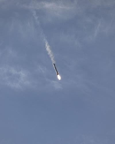 První stupeň Falconu 9 padá zpět atmosférou s roztaženými mřížkovými kormidly a slabou kondenzační stopou na modré obloze.