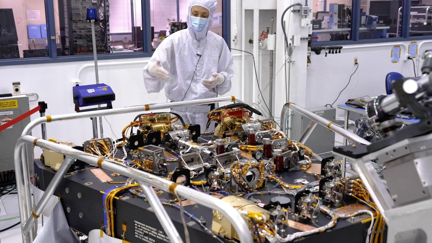 Technik v ochranném oděvu pracuje vedle optické sestavy koronografu Romanova teleskopu v čisté hale JPL.