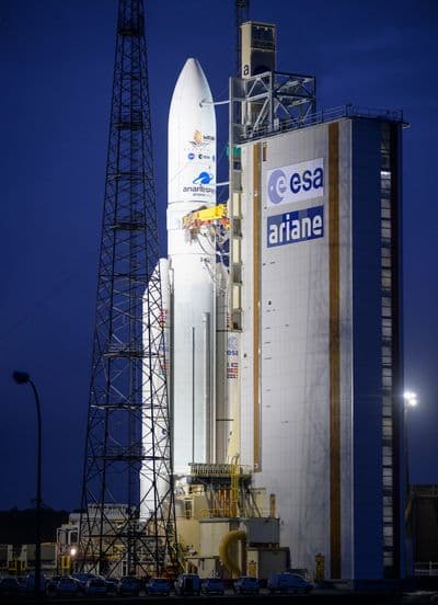 Raketa Ariane 5 s vesmírným dalekohledem Jamese Webba stojí za soumraku u obslužné věže s nápisy ESA a Ariane na kosmodromu Kourou.