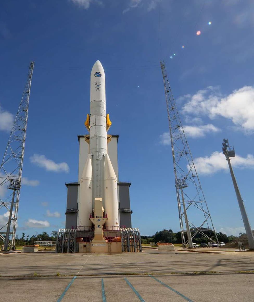 Evropská raketa Ariane 6 stojí na startovní rampě kosmodromu Kourou ve Francouzské Guyaně pod modrou oblohou, po stranách jsou bleskosvodné stožáry.